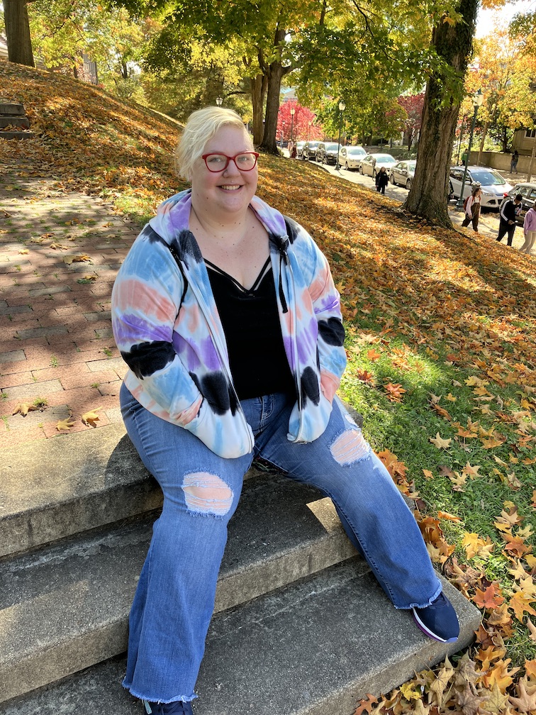 A smiling human with bright red glasses and short blonde hair, wearing a colorful tie dyed hoodie in pastels and ripped jeans.